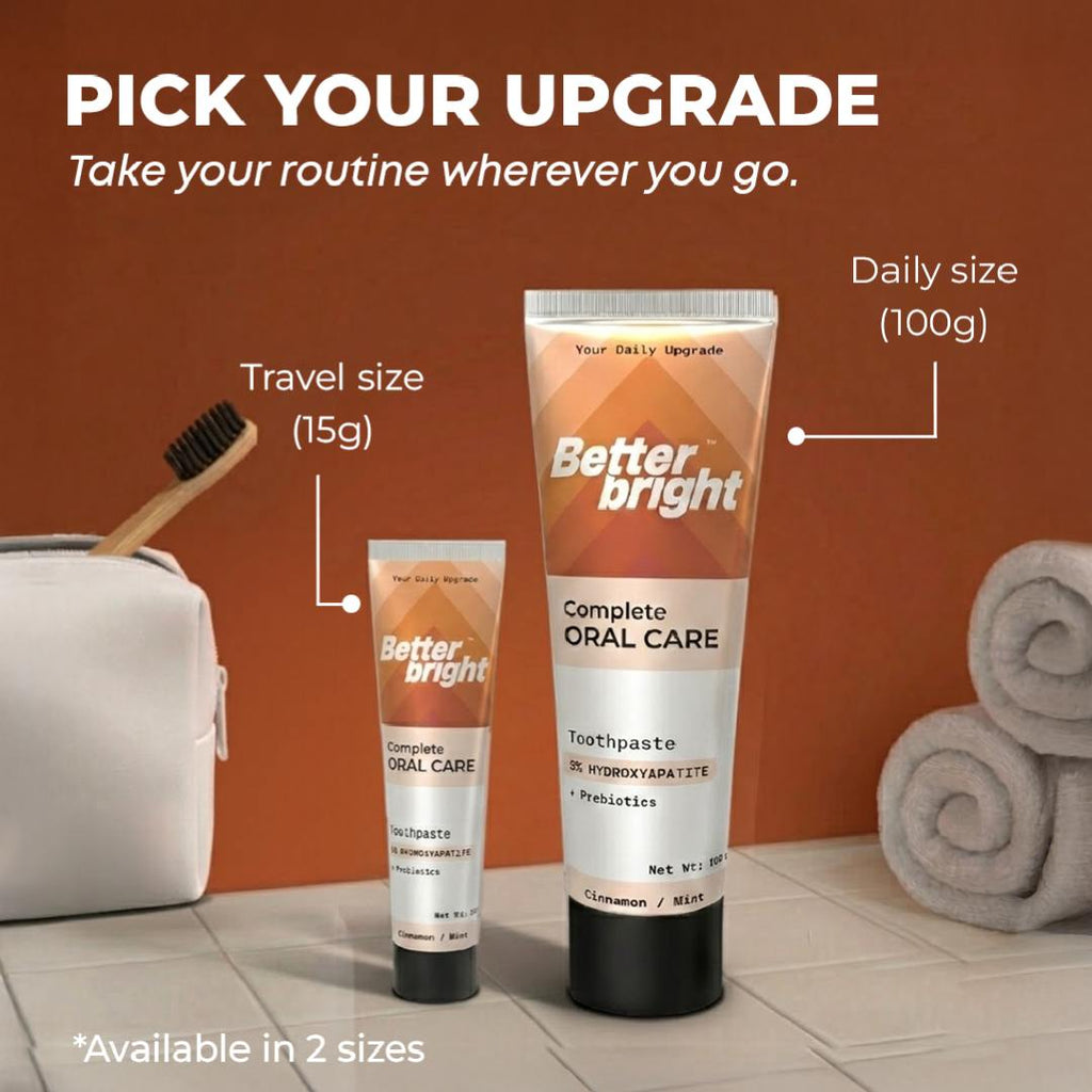 Two tubes of Better Bright Complete Oral Care Cinnamon mint toothpaste shown in travel size (15g) and daily size (100g), set against a warm brown background with bathroom props including a white pouch, toothbrush, and rolled towels.