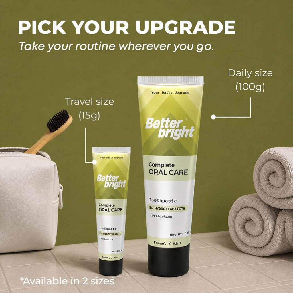 Two tubes of Better Bright Complete Oral Care fennel mint toothpaste shown in travel size (15g) and daily size (100g), set against a warm brown background with bathroom props including a white pouch, toothbrush, and rolled towels.
