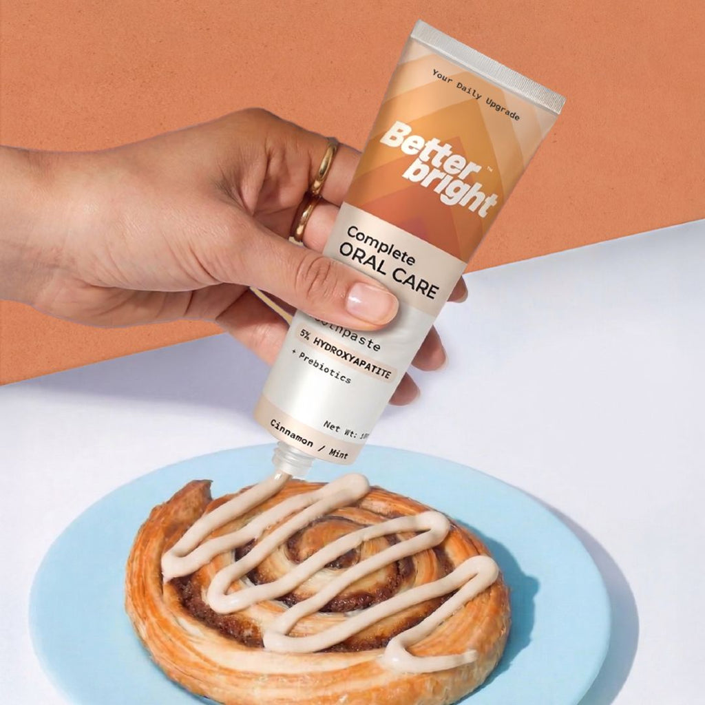 A hand holds a tube of Better Bright Complete Oral Care Cinnamon mint toothpaste above a cinnamon roll on a light blue plate, with the toothpaste being drizzled onto the pastry. The background is split diagonally between a warm peach color and a white surface.