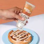 A hand holds a tube of Better Bright Complete Oral Care Cinnamon mint toothpaste above a cinnamon roll on a light blue plate, with the toothpaste being drizzled onto the pastry. The background is split diagonally between a warm peach color and a white surface.