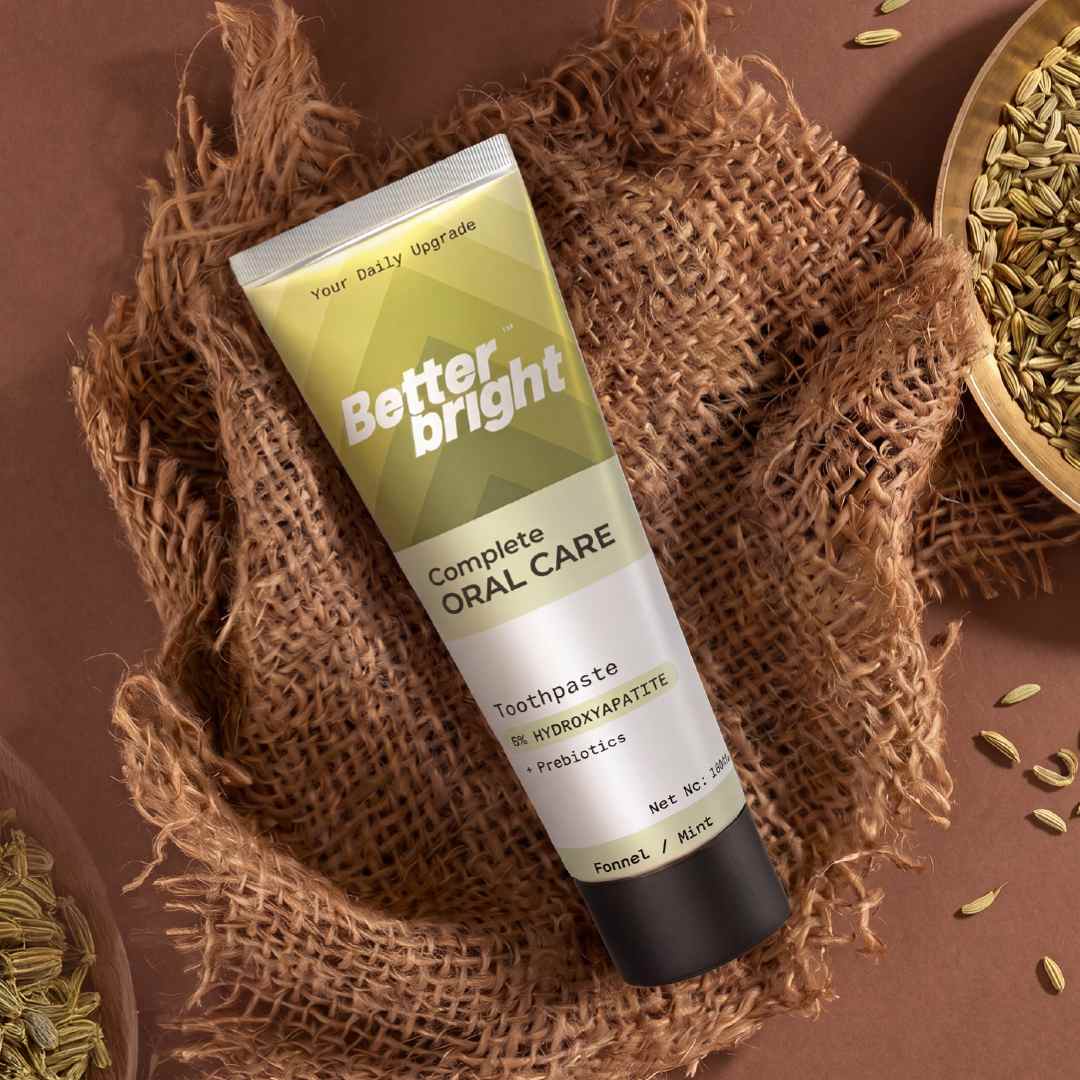 A tube of Better Bright Complete Oral Care Fennel mint toothpaste is displayed diagonally on a textured brown burlap cloth, surrounded by natural ingredients like fennel seeds in a wooden bowl.